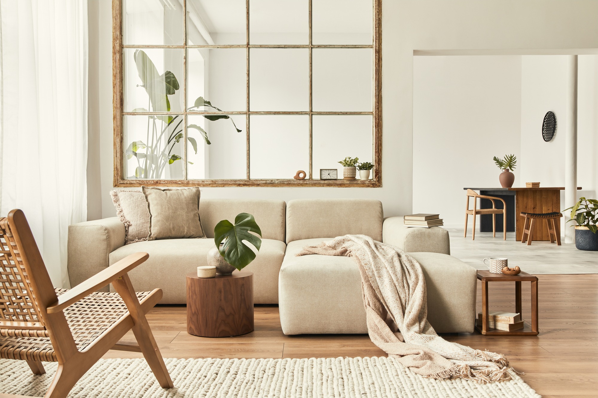 Cozy living room featuring a beige sectional sofa, a wooden coffee table, and a large rustic mirror. Lush plants enhance the inviting atmosphere.