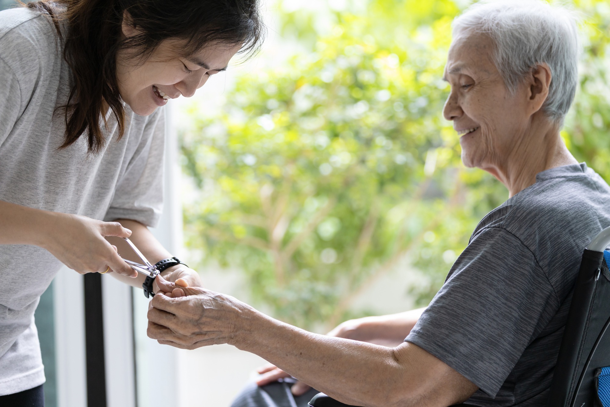 A caregiver gently trims the nails of an elderly person, both smiling, in a bright room with greenery in the background, highlighting compassion and care.