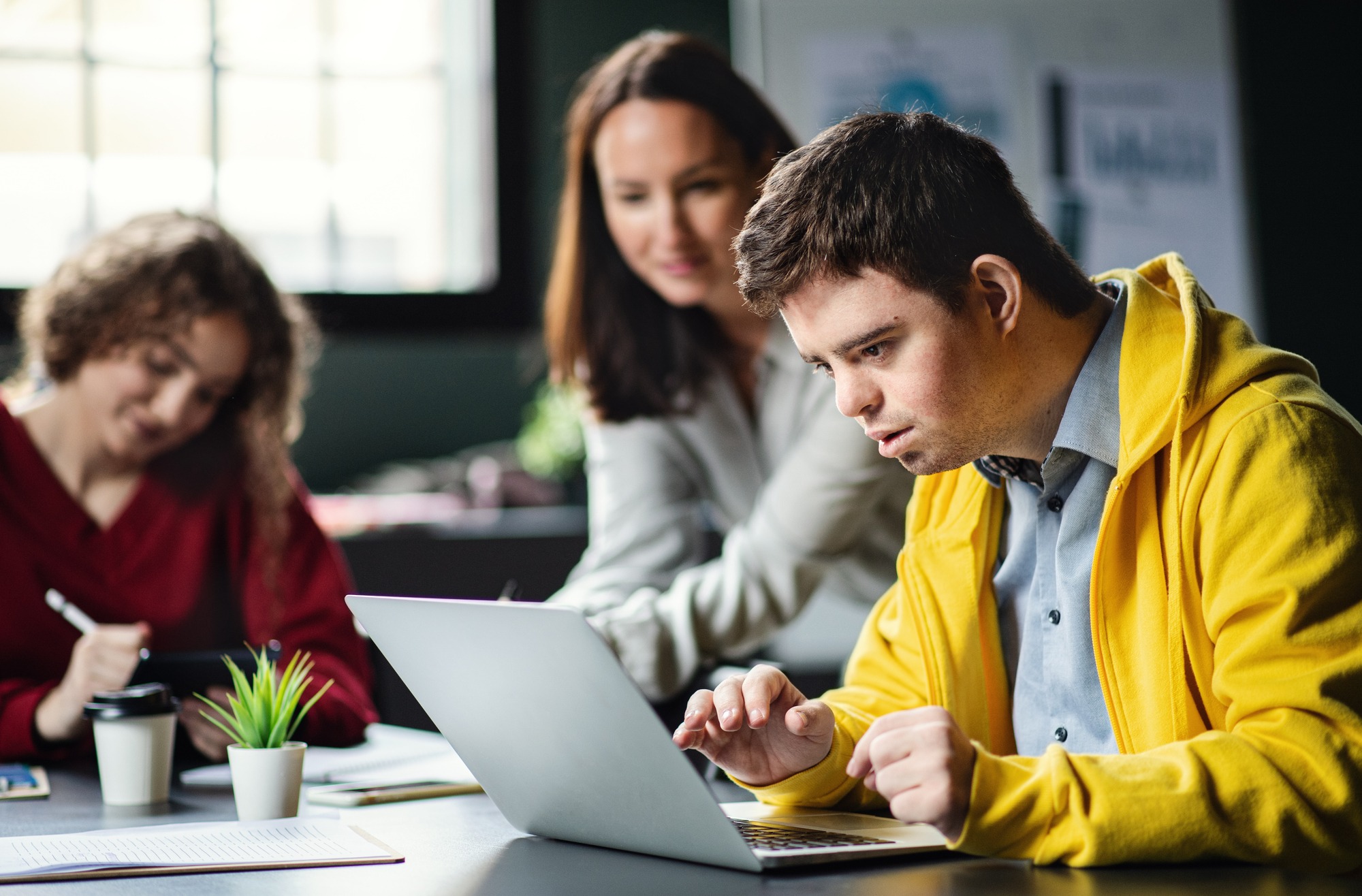 A young man in a yellow hoodie intently uses a laptop, assisted by a focused woman. Another woman notes in the background, highlighting collaboration in a learning environment.