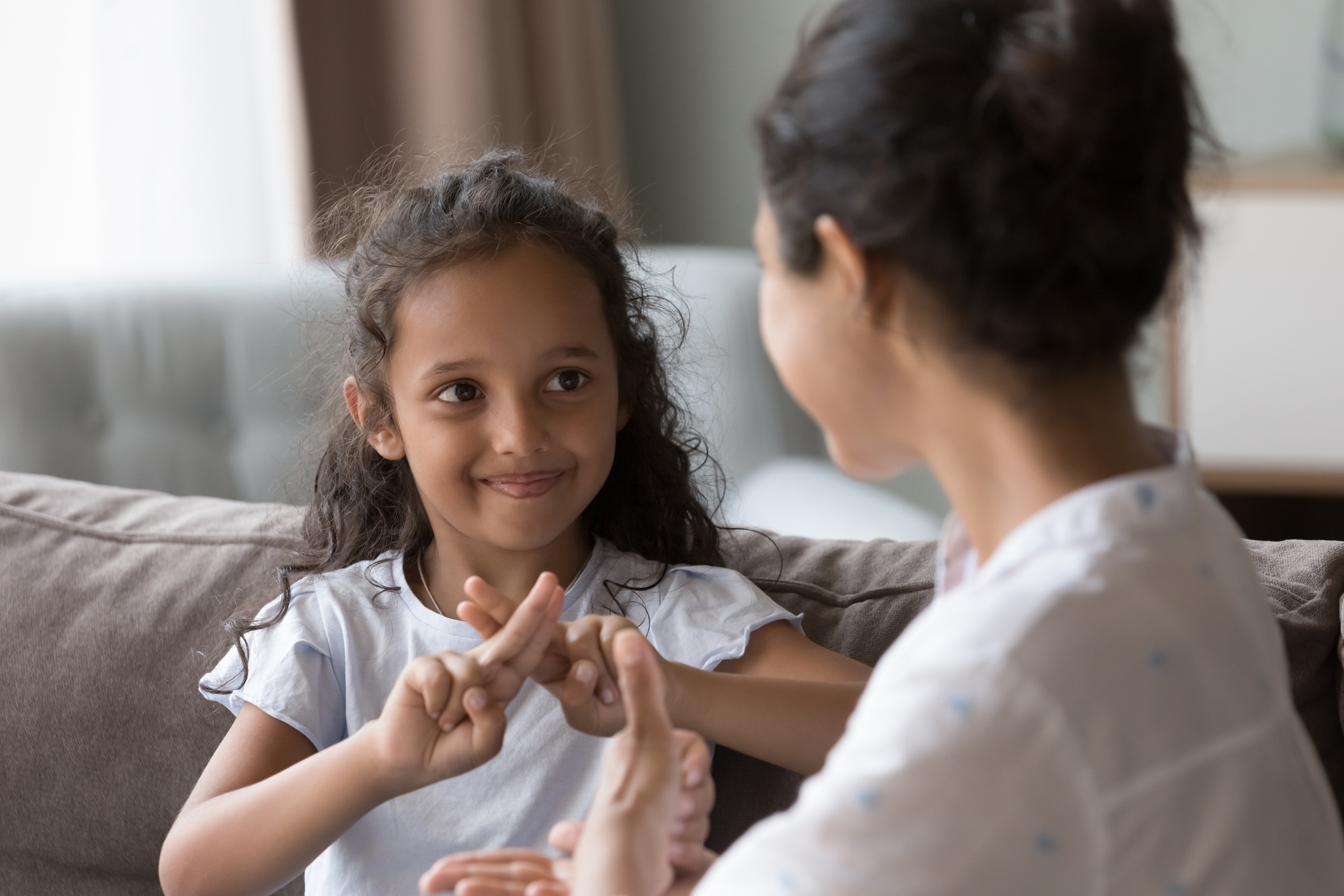 A young girl and an adult are engaged in a playful hand game, smiling and enjoying their interaction on a couch in a cozy living room.