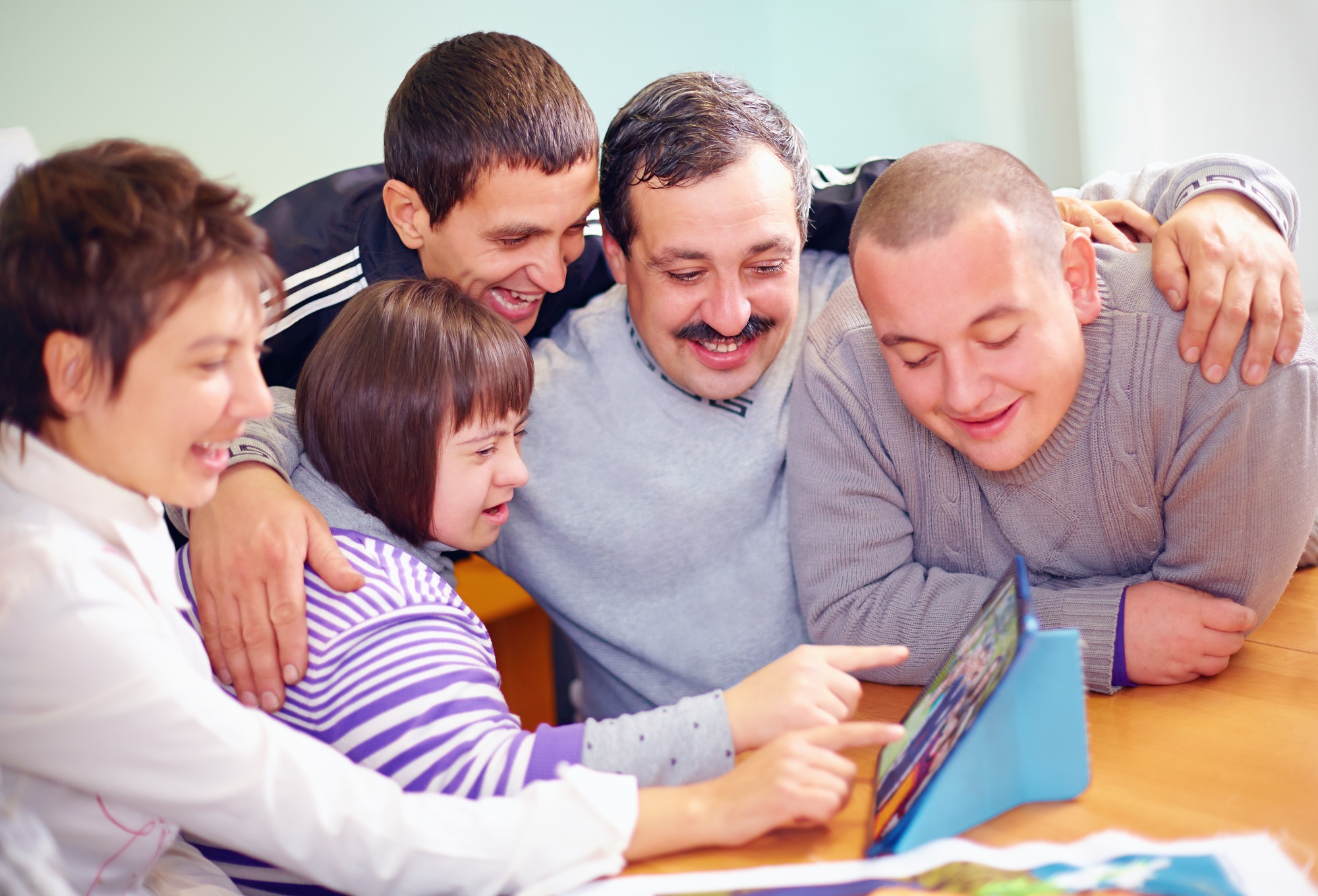 A joyful group of five, including a girl with Down syndrome, gathers around a tablet, sharing laughter and engaging in a game or activity together.